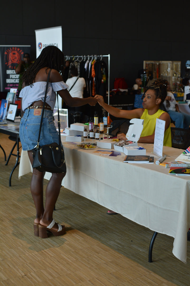 Two women shaking hands at a table with various items on display, possibly at an event or convention.