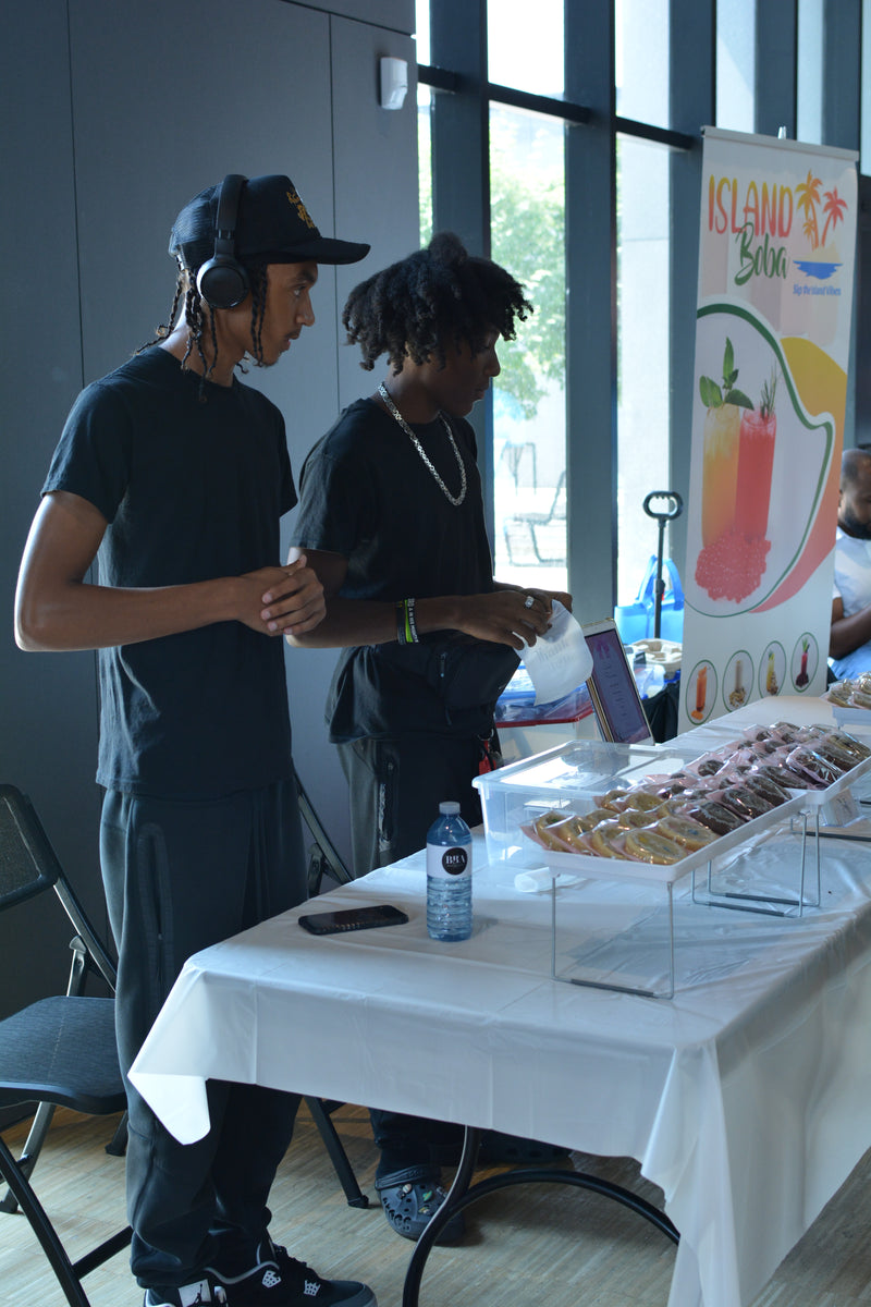 Two people standing near a table with donuts and a laptop, with a colorful sign in the background.