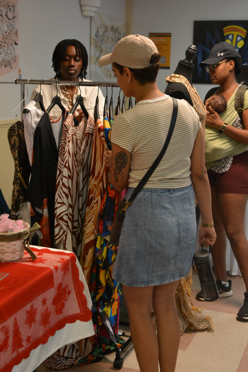 Woman looking at clothes on a rack with another person holding a baby in the background.