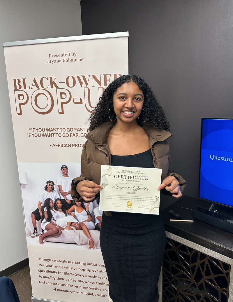 Woman holding a certificate in front of a 'Black-Owned Pop-Up' banner and computer screen.