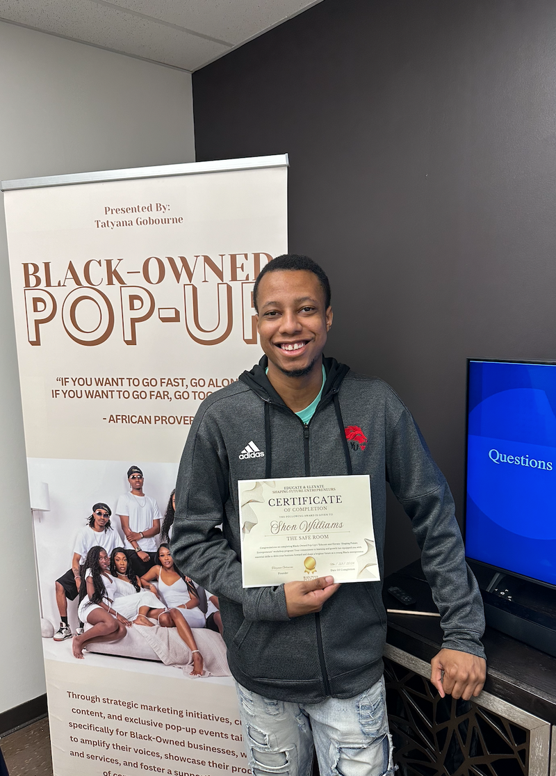Person holding a certificate in front of a 'Black-Owned Pop-Up' event banner.